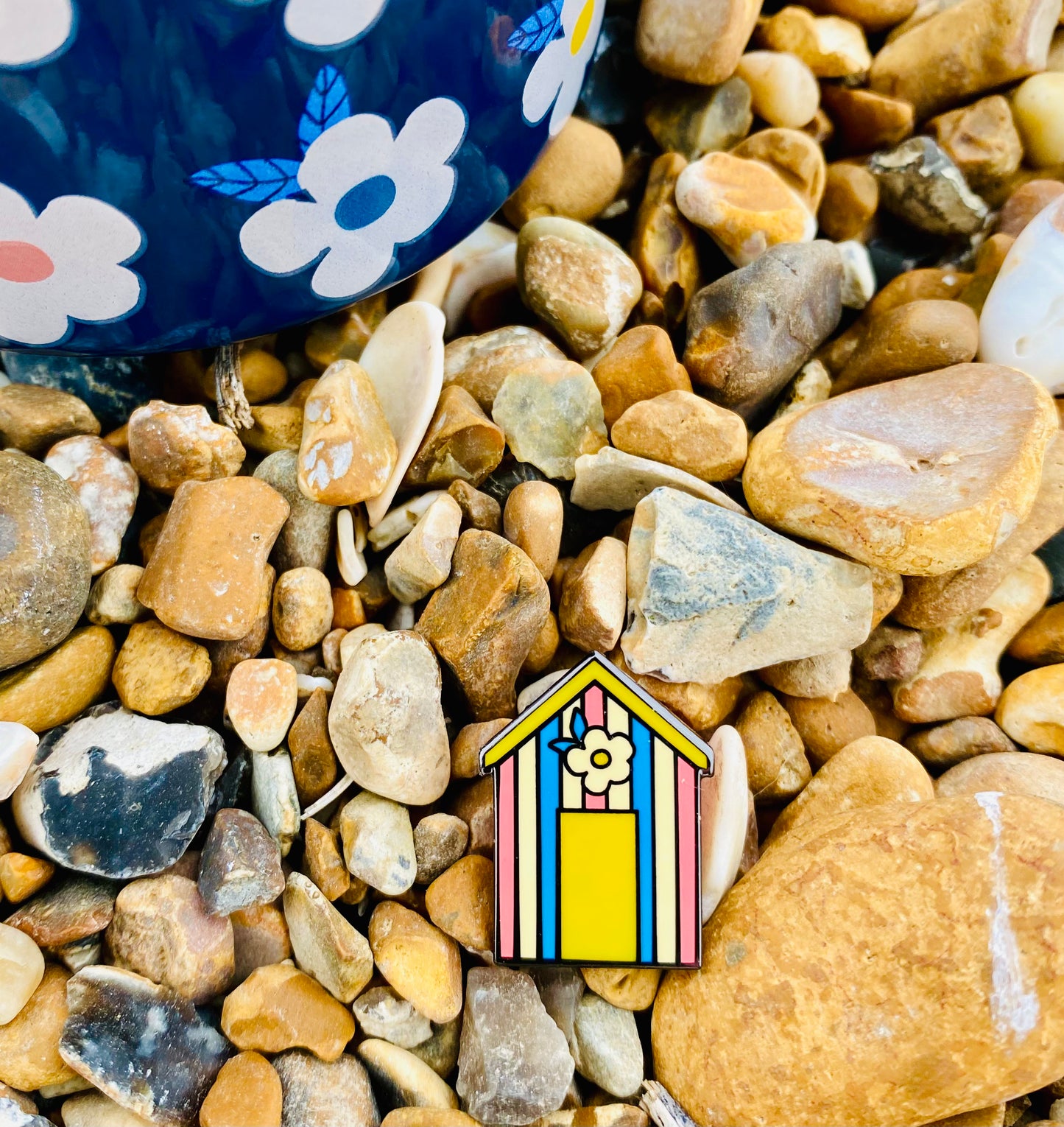 Cute candy stripe beach hut pin badge. The beach hut has pink, blue, yellow and cream stripes and features a yellow door and retro flower above. The photo was taken on the beach pebbles and also features our Enamelhappy midnight enamel mug