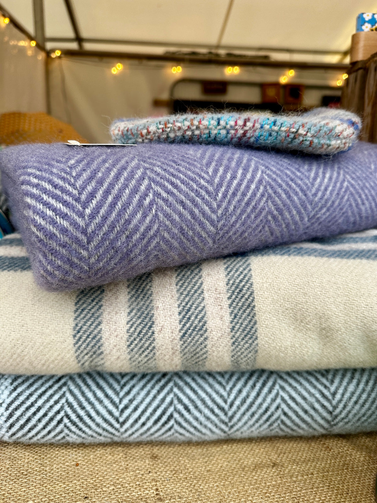 A stack of folded woven textiles, including a lavender-purple herringbone blanket, a cream and blue striped throw, and a light blue herringbone piece, with a small multicolored knit item on top, photographed in a tent-like setting.